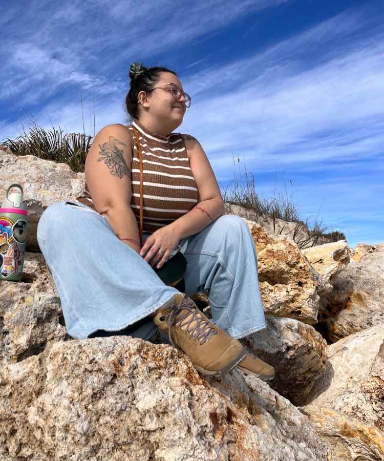 A imagem mostra uma mulher sentada em uma rocha sob um céu azul. Ela usa uma blusa listrada marrom, jeans largos e botas marrons. Seus cabelos estão presos com uma presilha verde. Ao lado, há uma garrafa decorada com adesivos coloridos.Influenciadoras plus size