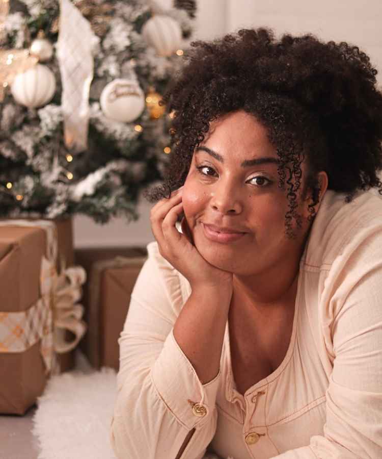 A imagem mostra uma mulher sorridente com cabelos cacheados e volumosos, vestindo uma blusa clara. Ela está deitada, apoiando o rosto na mão, em frente a uma árvore de Natal decorada com enfeites brancos e dourados, cercada por presentes embalados.
