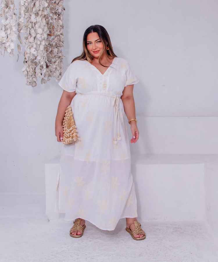 A imagem mostra uma mulher em um vestido branco longo com detalhes sutis. Ela segura uma bolsa de palha e usa sandálias douradas. Sua expressão é serena, e o cabelo está solto. O ambiente é minimalista com decoração floral ao lado.