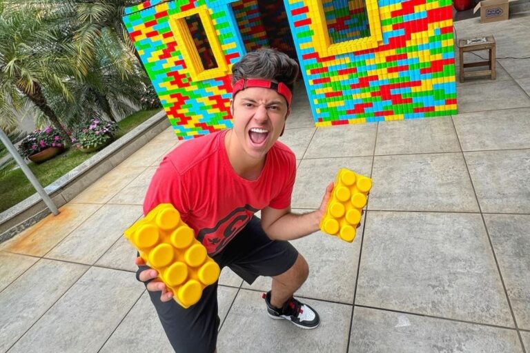 Enaldinho usando a blusa vermelha de sua marca, bermuda preta, segurando peças de Lego grandes e casa de Lego ao fundo