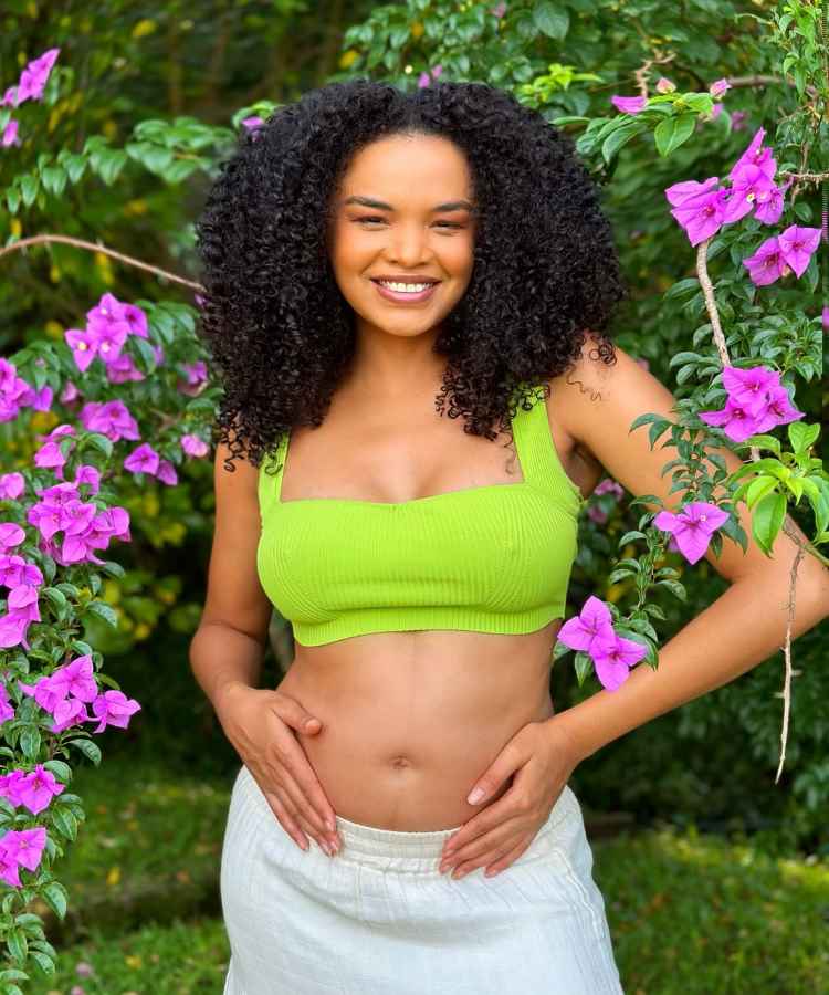 Uma mulher está sorrindo em meio a flores roxas e vegetação verde. Ela usa um top verde e saia branca, com as mãos repousando sobre a barriga. Seu cabelo é encaracolado e volumoso, e a cena transmite uma sensação de alegria e tranquilidade - famosas que anunciaram gravidez