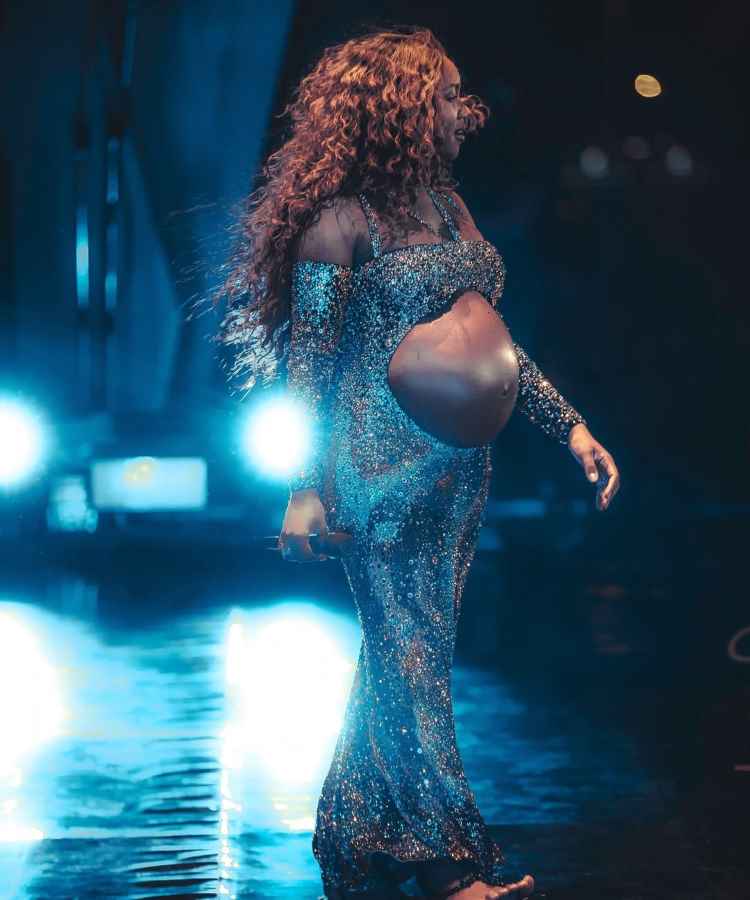 A imagem mostra uma mulher grávida em um palco iluminado. Ela veste um vestido longo e brilhante com um recorte que destaca a barriga. Seus cabelos são longos e cacheados. - famosas que anunciaram gravidez