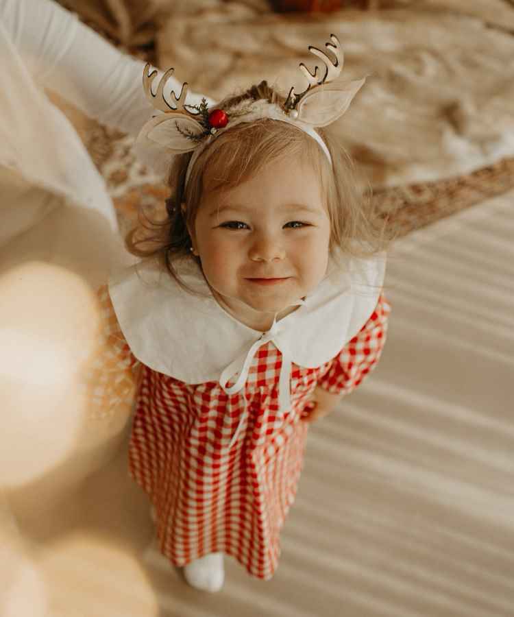 A imagem mostra uma menina sorridente, vestida com roupa xadrez vermelha e branca. Ela usa uma tiara com chifres de rena, decorada com uma pequena cereja. O ambiente é aconchegante, com luz suave destacando a expressão alegre da criança.