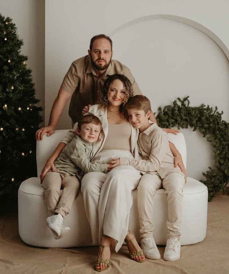 Família reunida em ambiente natalino, com árvore decorada ao lado. Mãe sentada em sofá branco, abraça dois filhos sorridentes. Pai inclinado atrás deles, todos vestidos em tons neutros. Fundo com guirlanda verde destaca a harmonia e alegria do momento festivo.