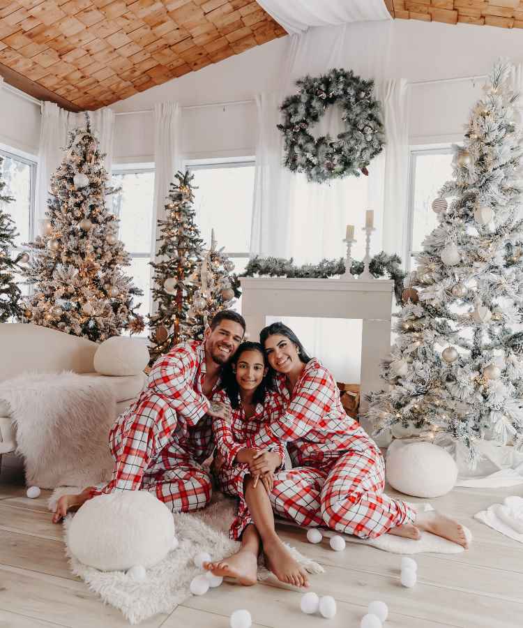 Família alegre em pijamas xadrez posa em sala decorada para o Natal, rodeada por árvores iluminadas e guirlanda. Estão sentados no chão, abraçados, transmitindo união e felicidade. O ambiente é acolhedor, com tons claros e detalhes festivos arredondados.