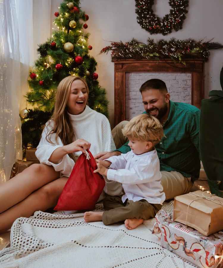 Família feliz sentada no chão, em ambiente natalino, decorado com árvore brilhante e guirlanda. Mulher segura saco de presente vermelho enquanto criança loira tenta abri-lo. Homem sorridente observa. Envolvidos em atmosfera aconchegante, cercados por luzes e embrulhos festivos.