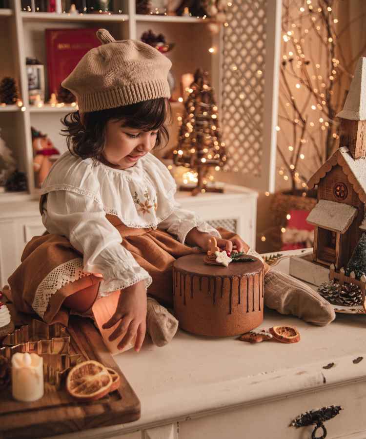 Menina sorridente sentada em uma mesa decorada para o Natal, com boina bege e vestido branco e marrom. Ela brinca com um bolo decorado e biscoito de gengibre. O ambiente é iluminado e aconchegante, com luzes e enfeites festivos ao fundo.