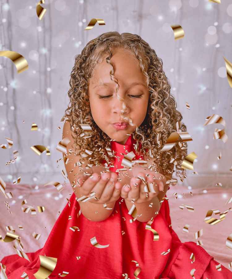 Menina de cabelos cacheados soprando confetes dourados, vestida com um vestido vermelho vibrante. O fundo é desfocado com luzes suaves, criando um efeito mágico e festivo. A cena transmite alegria e celebração, destacando o momento de encantamento e diversão.