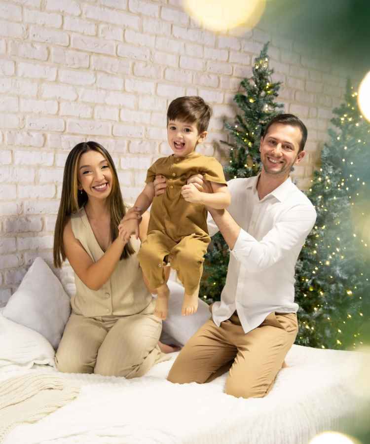 Família sorridente com pai, mãe e filho em ambiente natalino. Estão sentados em um sofá, com árvores de Natal decoradas ao fundo. O menino, feliz, é segurado pelos pais. A atmosfera é alegre e iluminada, transmitindo união festiva.