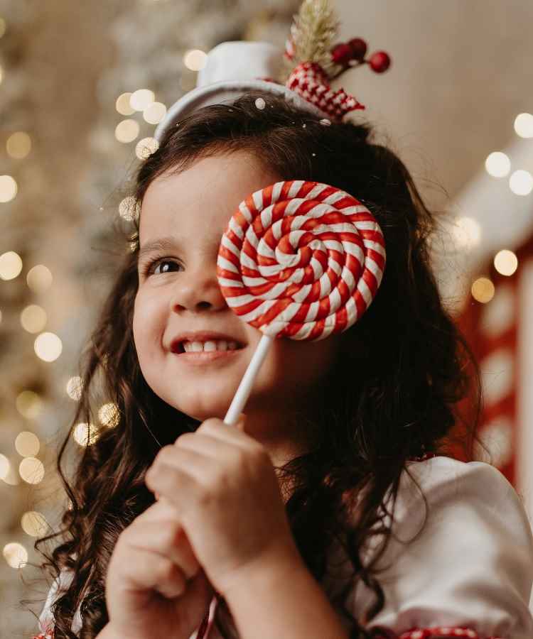 Menina sorridente com cabelo cacheado segura pirulito vermelho e branco, parcialmente cobrindo um olho. Ela usa tiara com enfeites natalinos. Ao fundo, luzes desfocadas criam uma atmosfera festiva e mágica, destacando a alegria do momento e o espírito natalino.