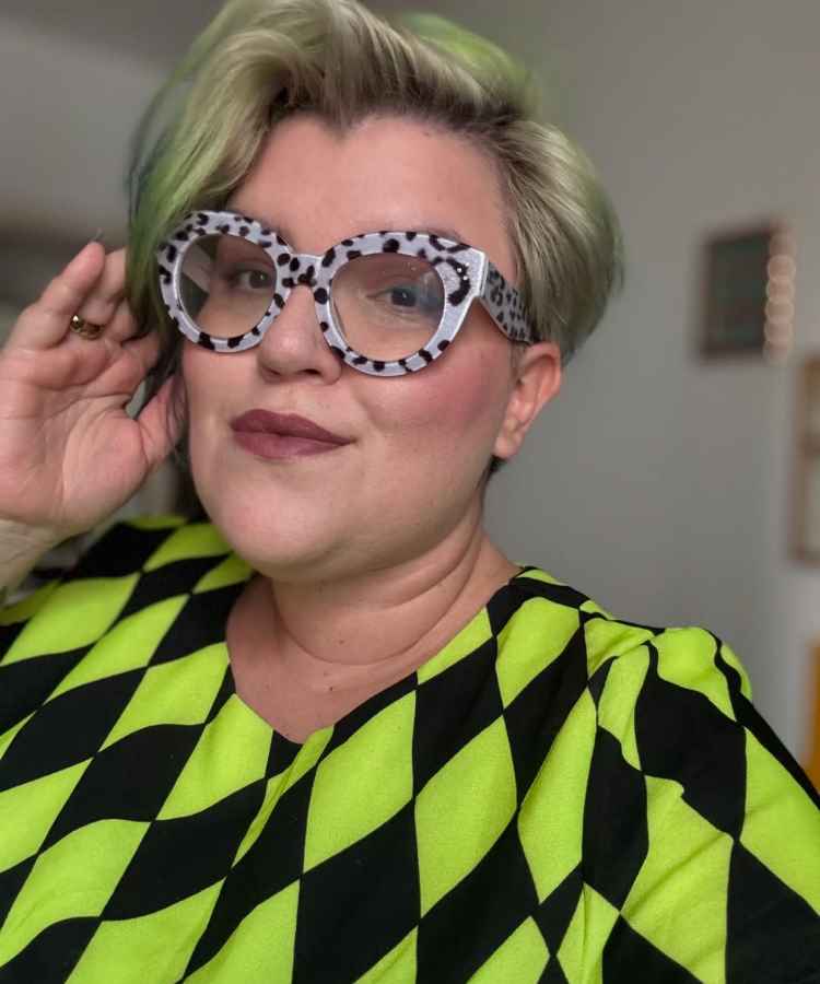 A imagem mostra uma mulher com cabelo loiro curto e óculos grandes de armação estampada. Ela veste uma blusa com padrão geométrico verde e preto. Sua expressão é confiante e estilosa, destacando um visual moderno e arrojado em um ambiente interno.