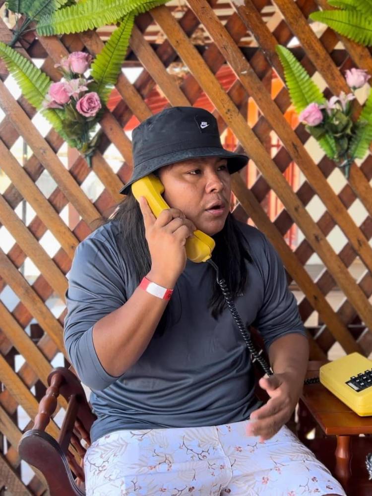 Kauri usando bucket hut cinza, blusa cinza e bermuda estampada, sentado em cadeira e com telefone amarelo ao ouvido