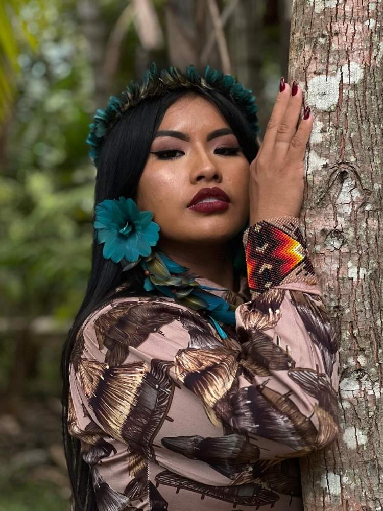 Mari Wapichana usando brinco de flor azul, pulseiras coloridas, vestido marrom, abraçada com tronco de árvore