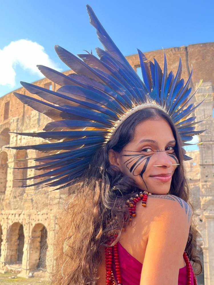 Alice Pataxó usando cocar azul, pintura no rosto e vestido vermelho em frente ao Coliseu