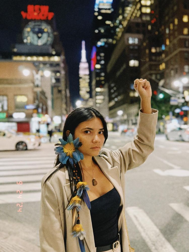 Priscila Tapajowara usando brinco de flor, roupa preta e blazer bege, fazendo símbolo de resistência na rua em Nova York