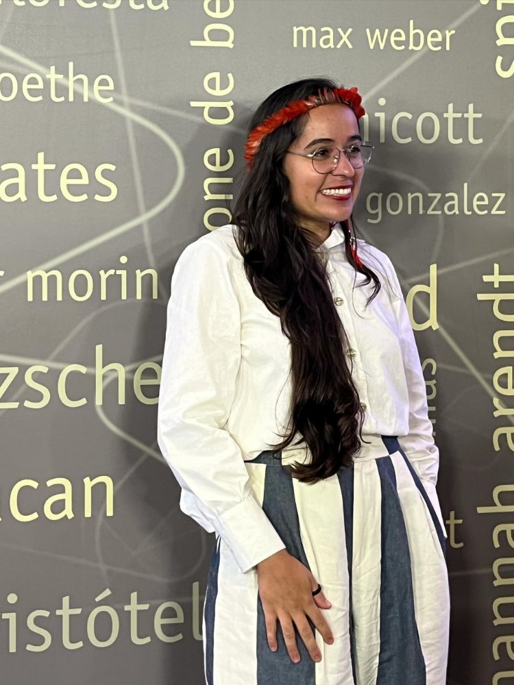 Geni Núñez usando roupa branca em frente a um mural cinza com nome de autores