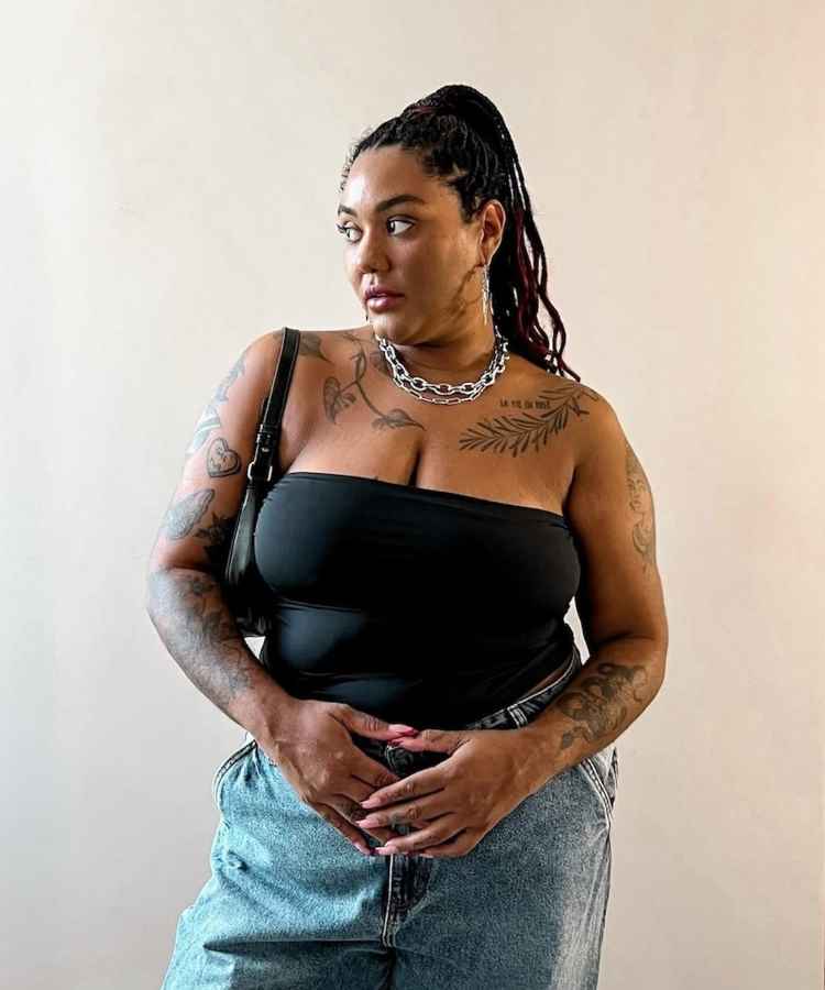 A imagem mostra uma mulher confiante vestindo um top preto sem alças e jeans. Ela tem tatuagens nos braços e ombros, além de uma corrente prateada no pescoço. Seu cabelo está preso em um coque com tranças.