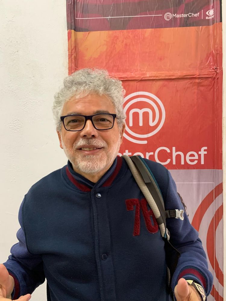 Dimas Souza usando jaqueta azul com logo vermelho do MasterChef ao fundo