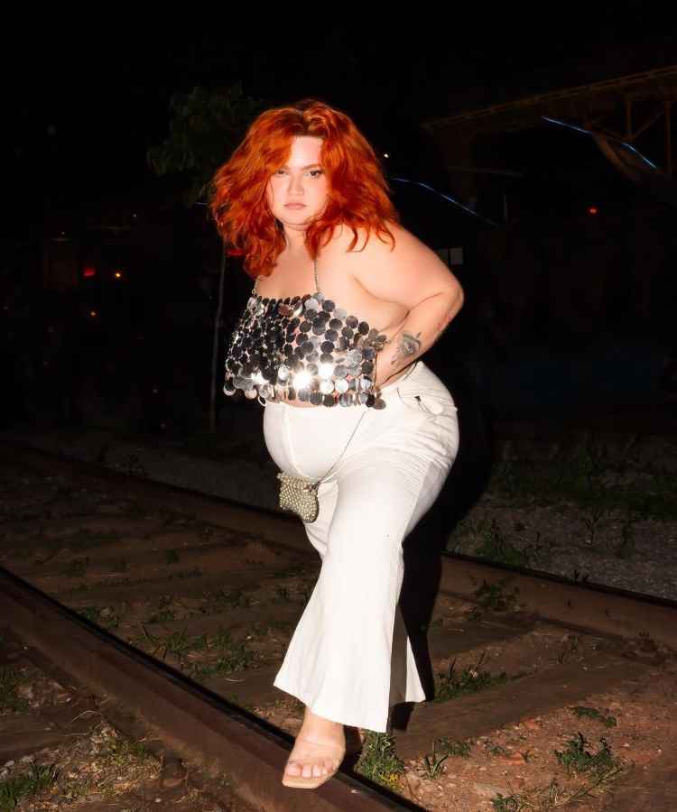 A imagem mostra uma mulher de cabelos ruivos e volumosos, em uma pose confiante. Ela usa um top brilhante de lantejoulas prateadas e calças brancas. Está em uma linha de trem à noite, destacando-se contra o fundo escuro.Influenciadoras plus size