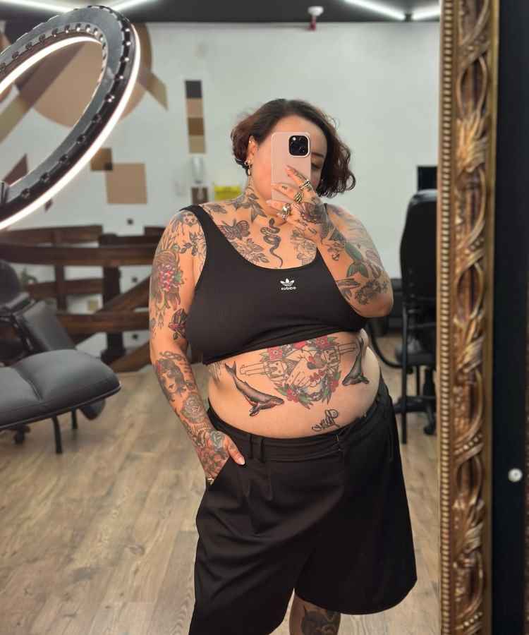 A imagem mostra uma mulher em um estúdio, tirando uma selfie. Ela veste um top preto Adidas e calças pretas. Seu corpo está coberto por tatuagens coloridas nos braços e barriga. O ambiente tem piso de madeira e um grande espelho.