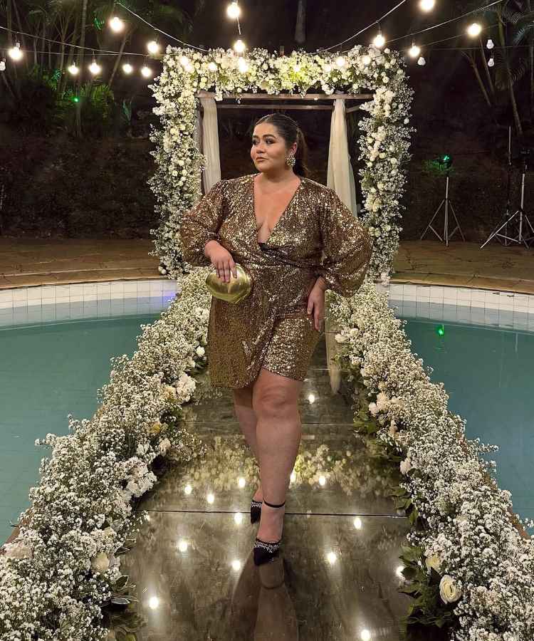 A imagem apresenta uma mulher em um vestido dourado cintilante, posando em uma passarela cercada por flores brancas. Ela segura uma bolsa metálica e está em um cenário noturno iluminado por lâmpadas suspensas, transmitindo elegância e sofisticação.