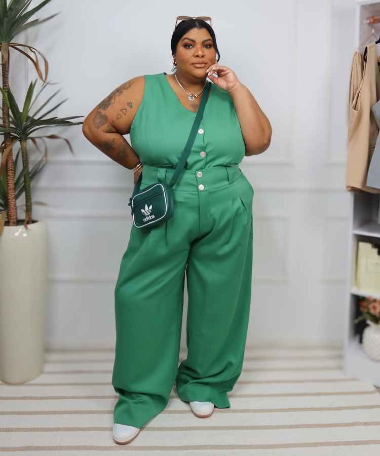 A imagem mostra uma mulher confiante usando um conjunto verde de calça e blusa sem mangas. Ela carrega uma bolsa Adidas verde. Com acessórios discretos, óculos de sol na cabeça e tatuagens no braço, ela exala estilo e atitude em um ambiente moderno.