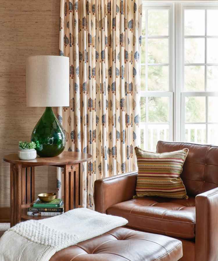 Sala de estar aconchegante com poltrona de couro e pufe combinando. Cortinas estampadas em tons neutros adicionam charme. Um abajur com base verde decora a mesa lateral ao lado de uma janela iluminada. Almofada listrada complementa o espaço acolhedor.