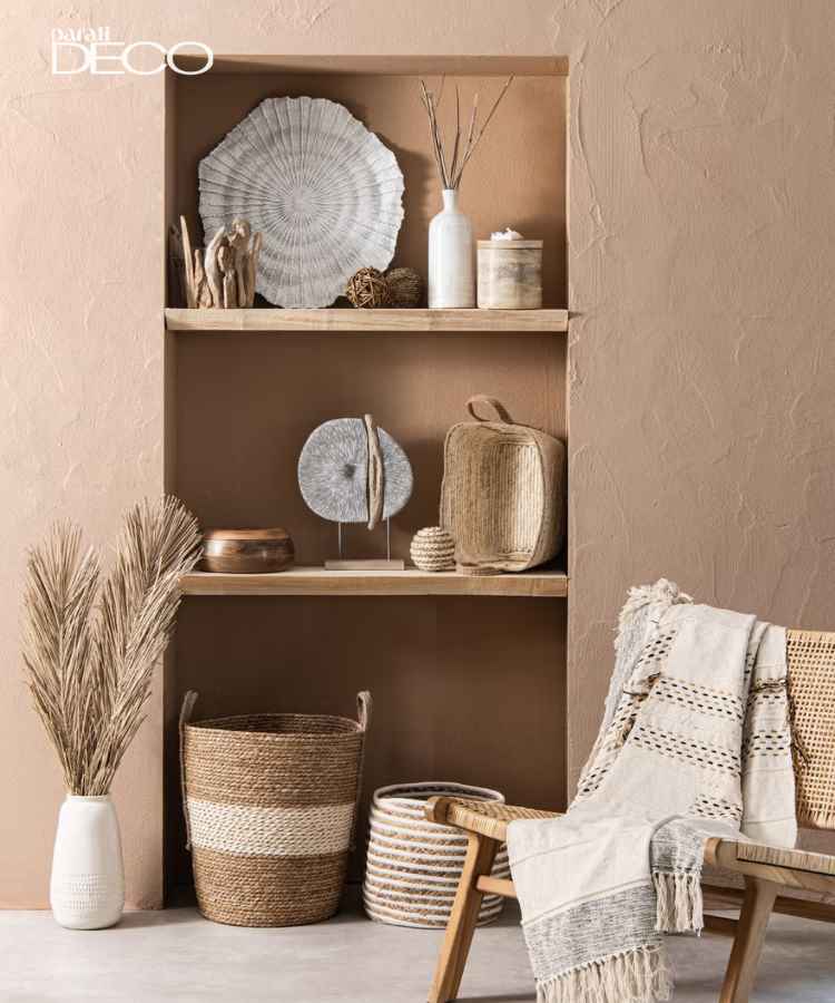 Ambiente rústico com prateleiras adornadas por objetos decorativos em tons neutros. Cestos de palha, vasos de cerâmica e esculturas naturais decoram o espaço. Uma cadeira com manta texturizada complementa a estética terrosa e acolhedora, destacando o equilíbrio entre forma e função.