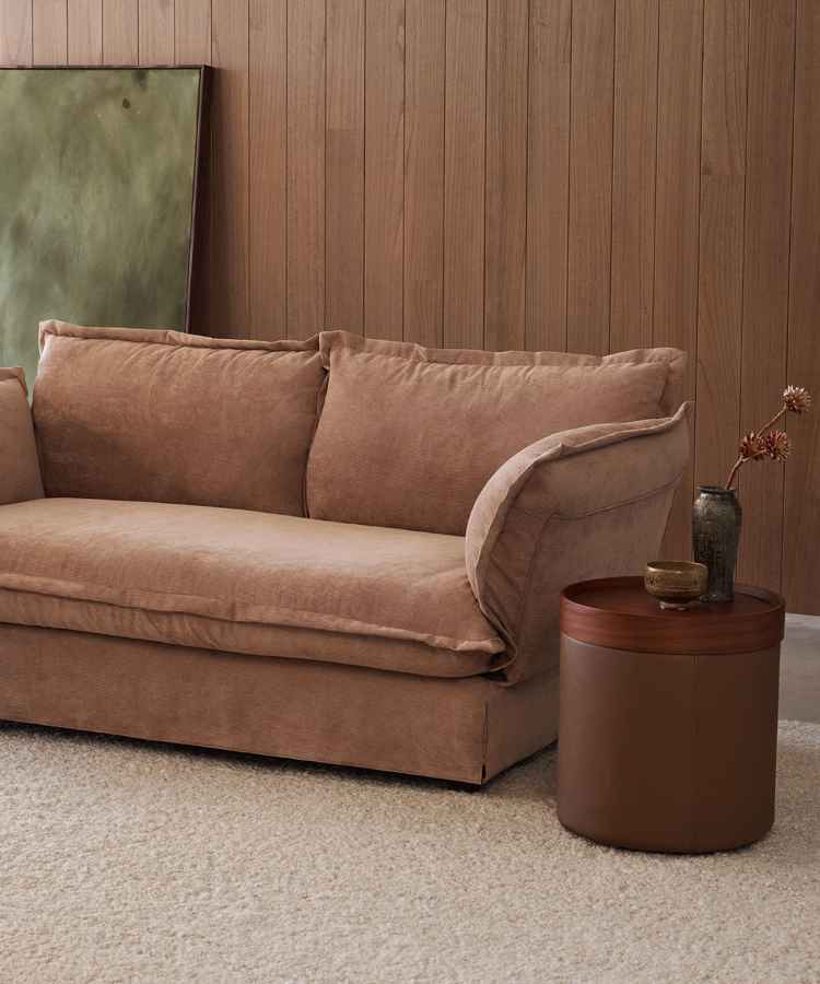 Ambiente acolhedor com sofá Mocha Mousse de tecido macio, complementado por uma mesa lateral redonda de madeira. Parede de madeira adiciona um toque rústico. Decoração minimalista inclui vasos simples sobre a mesa, criando uma atmosfera tranquila e elegante.