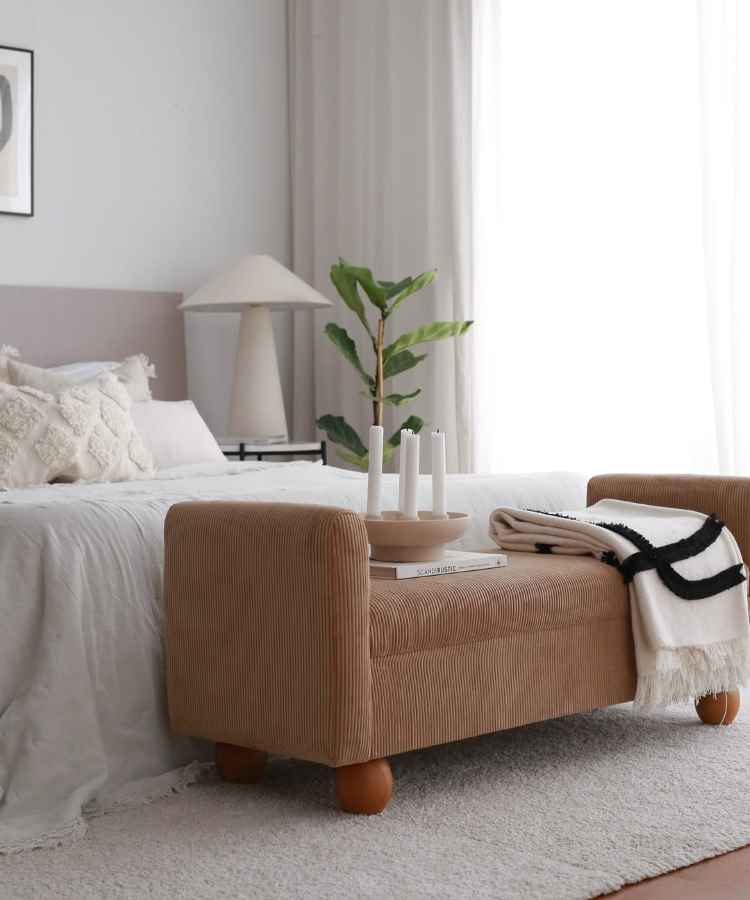 Quarto elegante com cama de linho branco e almofadas texturizadas. Um banco estofado em veludo marrom claro está aos pés da cama, decorado com livros e velas. Ao fundo, uma luminária moderna e planta verde complementam o ambiente sereno e sofisticado.