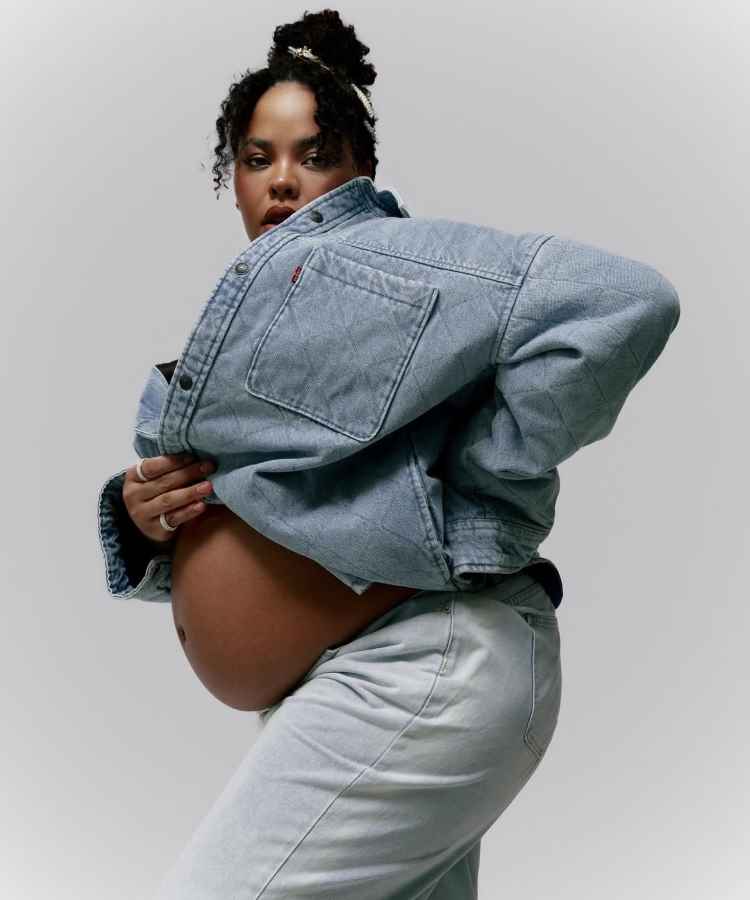 A imagem mostra uma mulher grávida em um casaco jeans azul claro, levemente aberto, revelando sua barriga arredondada. Ela exala confiança e força, com um olhar determinado. O cabelo está preso em um coque despojado, criando um visual moderno e impactante.