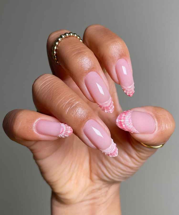 Mão com unhas decoradas em tom rosa claro com pontas texturizadas em rosa e branco. As unhas têm formato oval e comprimento médio. O fundo é cinza. A mão usa anéis dourados, criando um efeito elegante e moderno no visual.