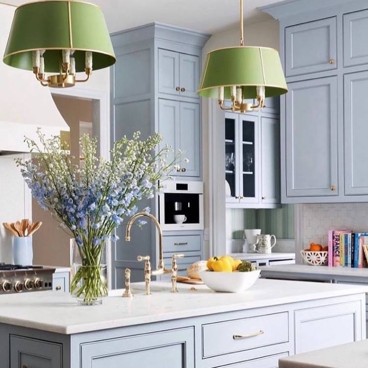 A imagem retrata uma cozinha sofisticada com armários azul-claro, luminárias verdes pendentes e detalhes dourados. Sobre a bancada branca, há um vaso com flores azuis e uma tigela com frutas. Livros coloridos decoram uma prateleira ao fundo.