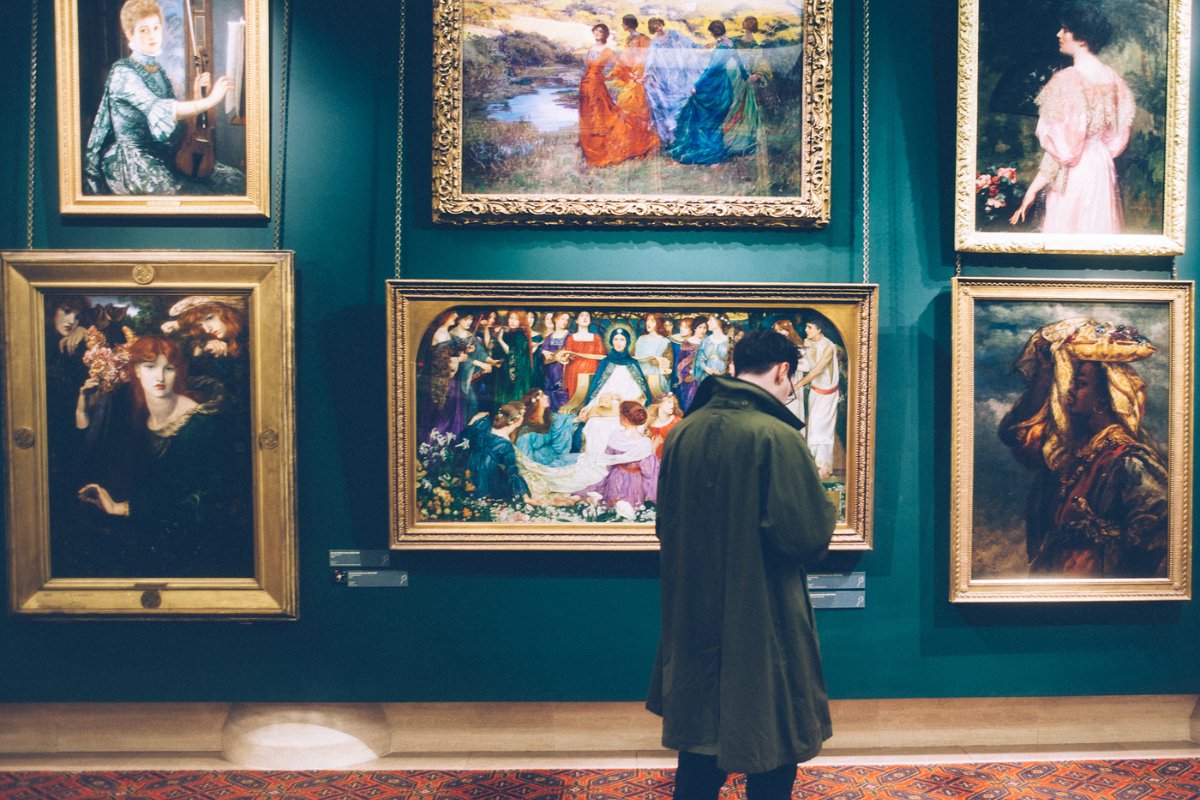 Museu com parede verde cheia de quadros e homem de pele clara com sobretudo escuro