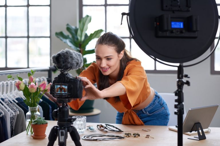Mulher de pele clara usando blusa laranja e calça jeans, ajeitando a câmera ao lado de ring light