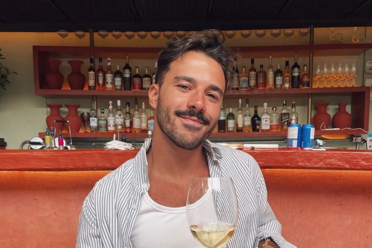 Mateus Pires usando regata branca e camisa azul e tomando vinho
