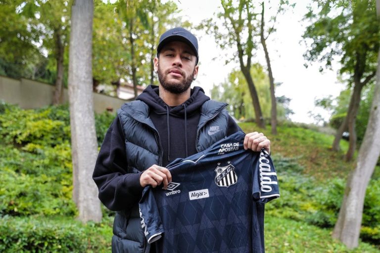 Neymar em jardim usando moletom preto, colete puffer azul, boné e segurando camisa azul do Santos