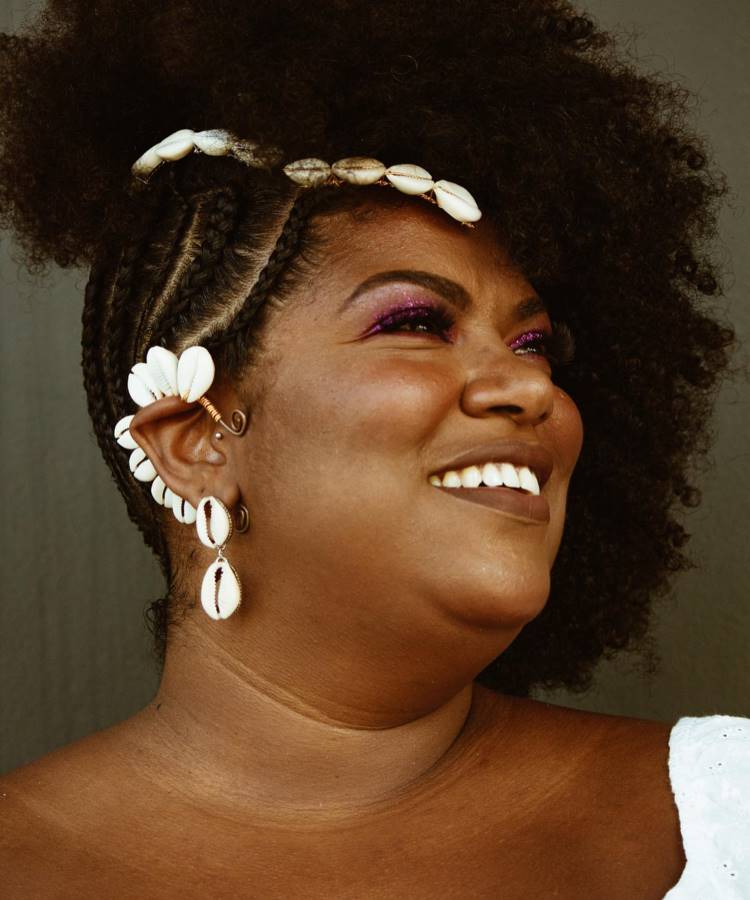 A mulher sorri radiante, exibindo penteado afro com tranças decoradas por conchas e um volumoso cabelo natural. Ela usa maquiagem com sombra rosa brilhante, brincos de conchas e um acessório de orelha, criando um visual elegante e autêntico.