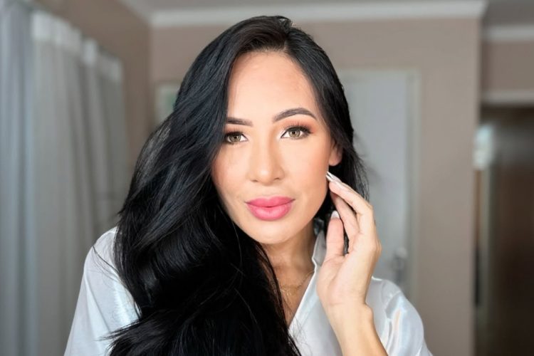 A imagem mostra Arleane Marques com longos cabelos pretos, pele radiante, maquiagem elegante e lábios levemente rosados. Ela usa uma blusa clara e olha diretamente para a câmera, segurando delicadamente o rosto. O fundo é neutro e desfocado.