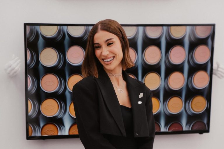 Boca Rosa de terno preto sorrindo, com foto de seus tons de base ao fundo