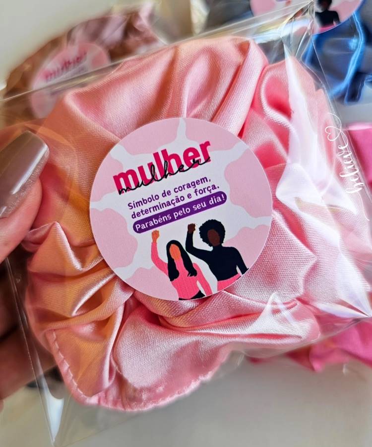 Lembrancinha com scrunchie de cetim rosa, embalada em plástico transparente, destacada por adesivo com a mensagem: "Mulher, símbolo de coragem, determinação e força. Parabéns pelo seu dia!" Fundo neutro com foco em estética feminina e comemorativa.