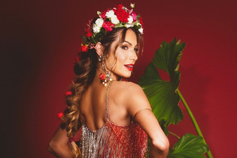 Mulher em pose elegante com vestido brilhante, coroa de flores vermelhas e brancas, cabelo trançado decorado, maquiagem marcante e fundo vermelho. Ela transmite charme e celebração, evocando o espírito vibrante do Carnaval.
