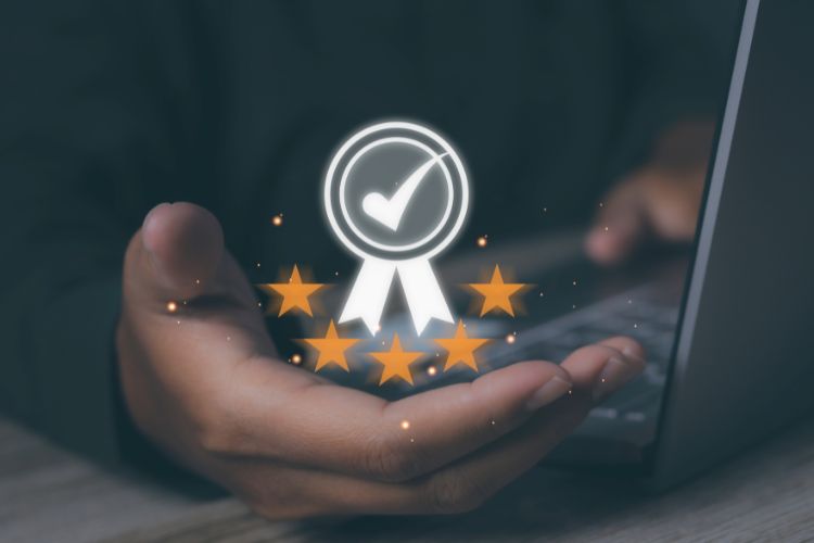Mão estendida segurando símbolo digital com selo de aprovação, contendo um check central e cinco estrelas douradas abaixo, representando qualidade, confiança e excelência, com fundo desfocado e um laptop ao lado para um contexto tecnológico.