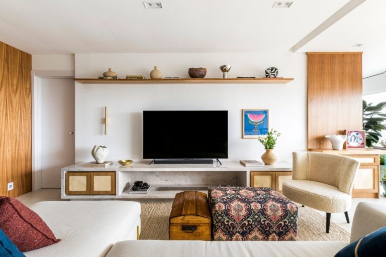 Sala de estar decorada com móveis sofisticados, incluindo painel de mármore branco Paraná, armários de madeira com palhinha, sofá claro, puff estampado, baú de madeira, prateleira com objetos decorativos e iluminação natural destacando o ambiente aconchegante e moderno.