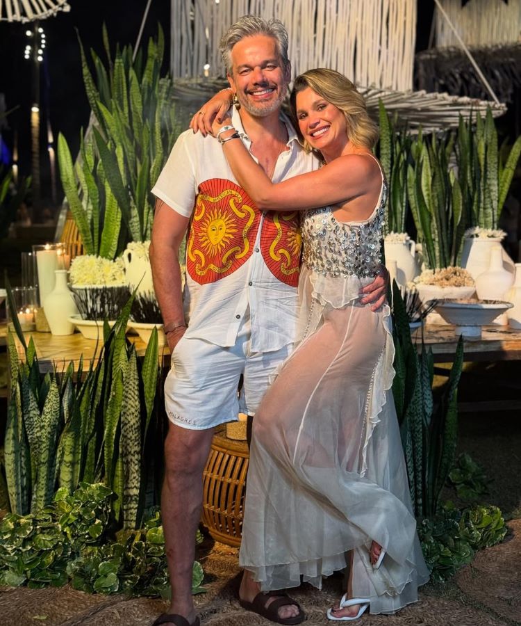 Flávia Alessandra e Otaviano Costa sorri abraçado em um ambiente decorado ao ar livre, rodeado por plantas. O homem veste camisa branca com detalhes vermelhos e shorts, enquanto a mulher usa vestido translúcido elegante, transmitindo alegria. Velas decoram o fundo iluminado.