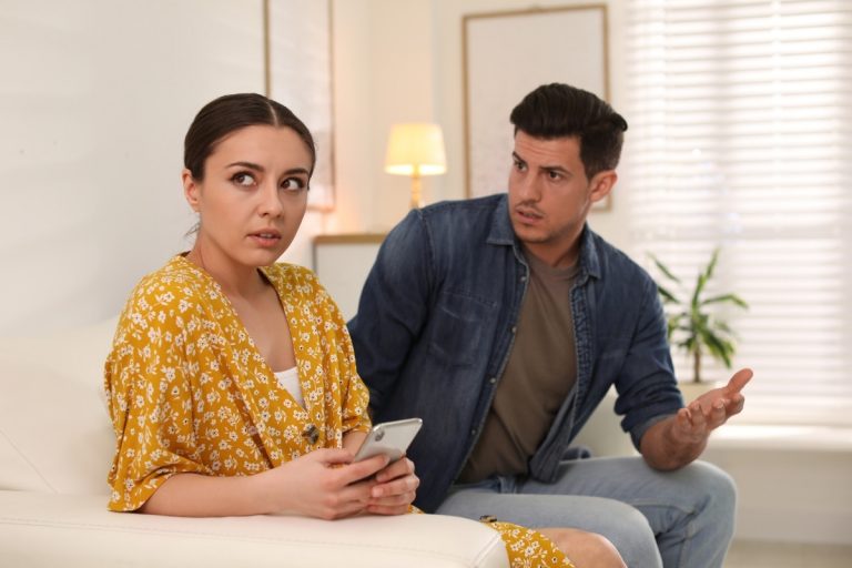 Uma mulher sentada em um sofá, segurando um celular, aparenta olhar desconfortável. Ao lado, um homem gesticula com expressão séria, sugerindo um momento de tensão ou discussão em um ambiente doméstico iluminado.