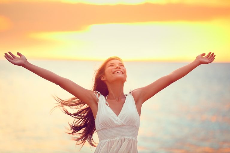 Mulher sorridente, de braços abertos, vestindo branco, aproveita o nascer ou pôr do sol à beira-mar. O céu em tons quentes de laranja e amarelo cria uma atmosfera serena e radiante, transmitindo liberdade e felicidade.
