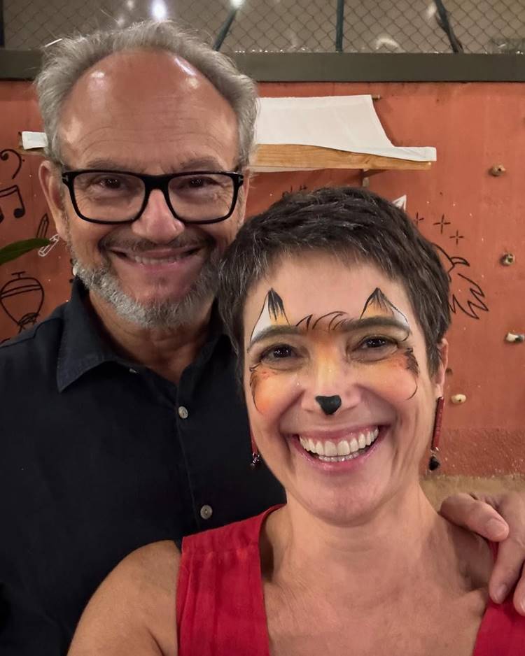 Um casal sorridente posa para a foto. O homem usa óculos e camisa azul escura, enquanto a mulher, com maquiagem de raposa e vestido vermelho, está à frente. Fundo apresenta uma parede laranja com desenhos animados e detalhes decorativos.