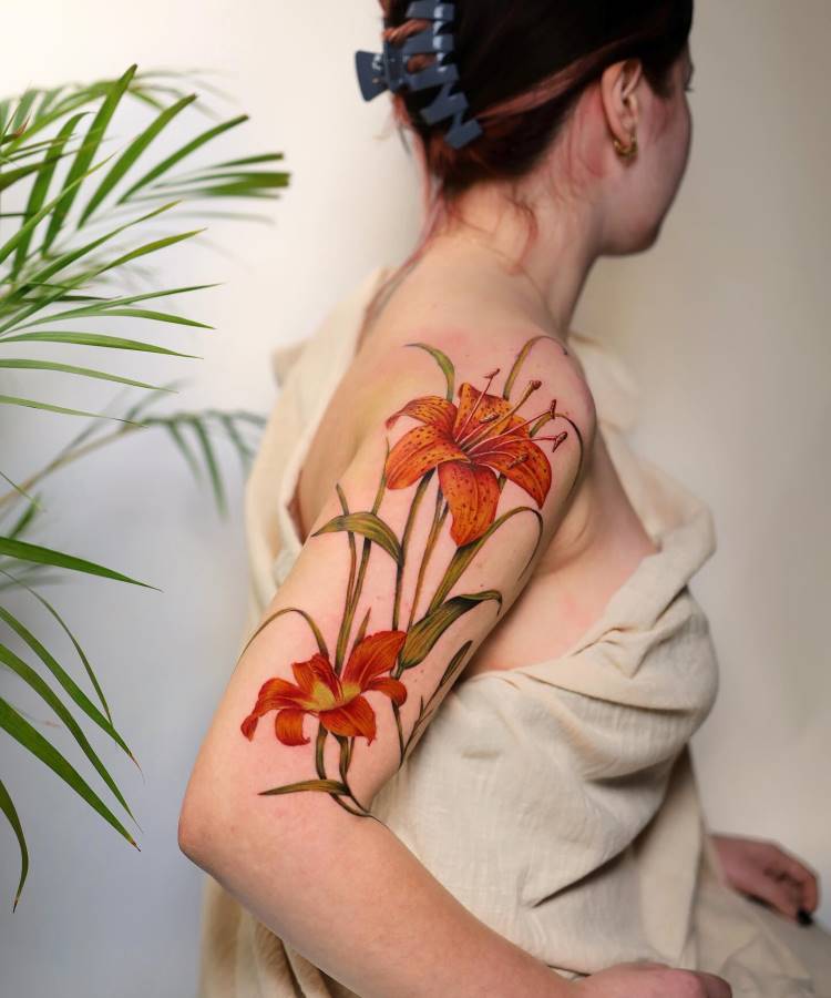 Tatuagem detalhada de flores laranjas com hastes verdes no braço de uma pessoa. As pétalas possuem tons vibrantes e realistas. A pele está coberta parcialmente com um tecido bege, e há plantas ao fundo desfocado.