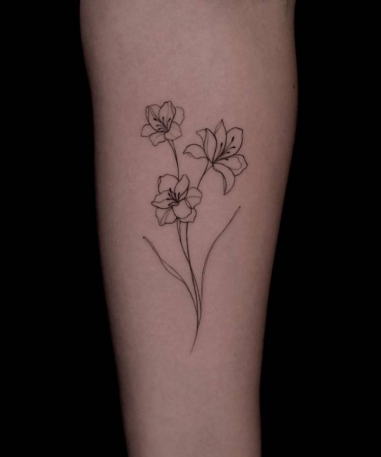 Tatuagem minimalista no braço com três flores delicadas desenhadas em linhas pretas finas. O design inclui hastes suaves e pétalas detalhadas, criando um visual elegante. Fundo preto destaca os traços da tatuagem, visível sobre a pele clara.